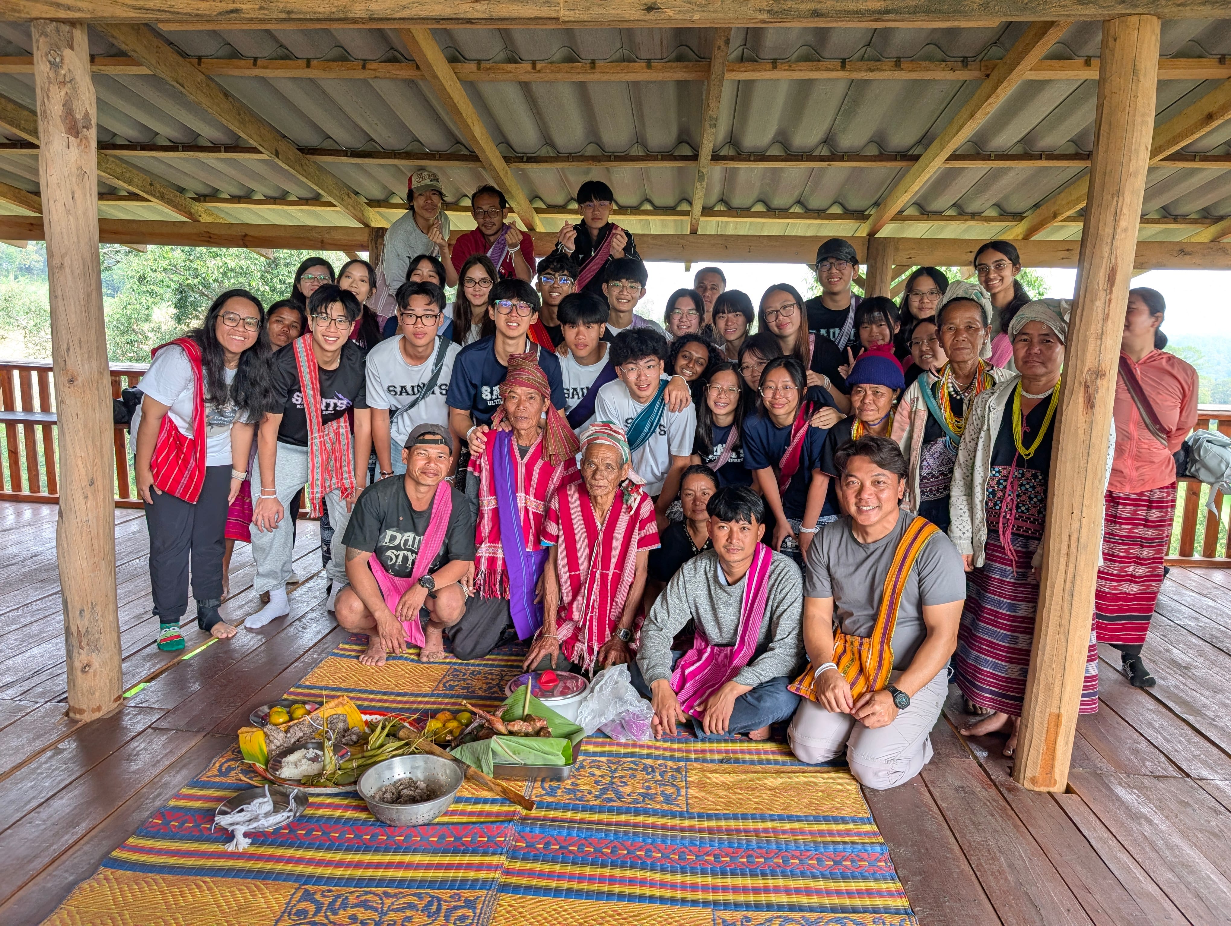 A group photo of student volunteers and locals sitting together inside a wooden shelter, gathered around traditional food spreads on woven floor mats.